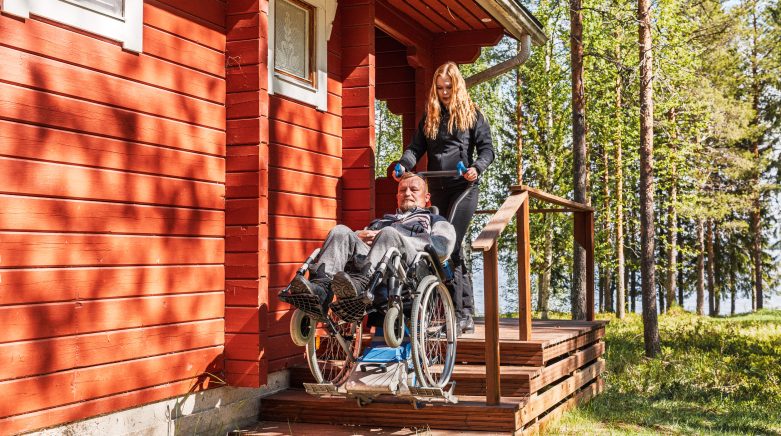Kuljettaja avustaa py&ouml;r&auml;tuolilla kuistin portaissa